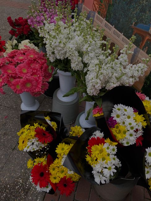 A display of fresh floral arrangements and bouquets arranged outdoors, featuring a variety of vibrant flowers including pink daisies, white stock, red roses, yellow chrysanthemums, and white daisies,