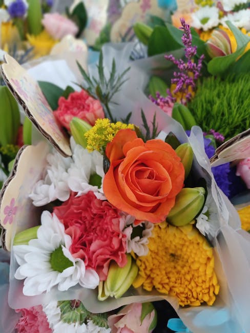 A vibrant floral arrangement featuring a large, soft orange rose at the center, surrounded by white daisies with green centers, pink carnations, yellow chrysanthemums, and small purple and yellow flow