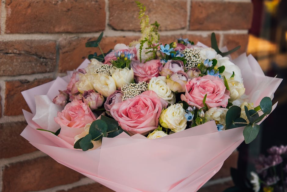 A bouquet of fresh pink roses, white lisianthus, and small blue and white flowers arranged with green eucalyptus leaves, wrapped in light pink tissue paper. The bouquet is held in a person's hand, aga