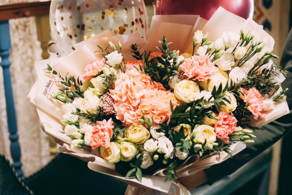 A large floral bouquet featuring soft pink carnations, white roses, and creamy ranunculus, accented with dark green foliage. The arrangement is wrapped in light pink paper with a subtle sheen and secu