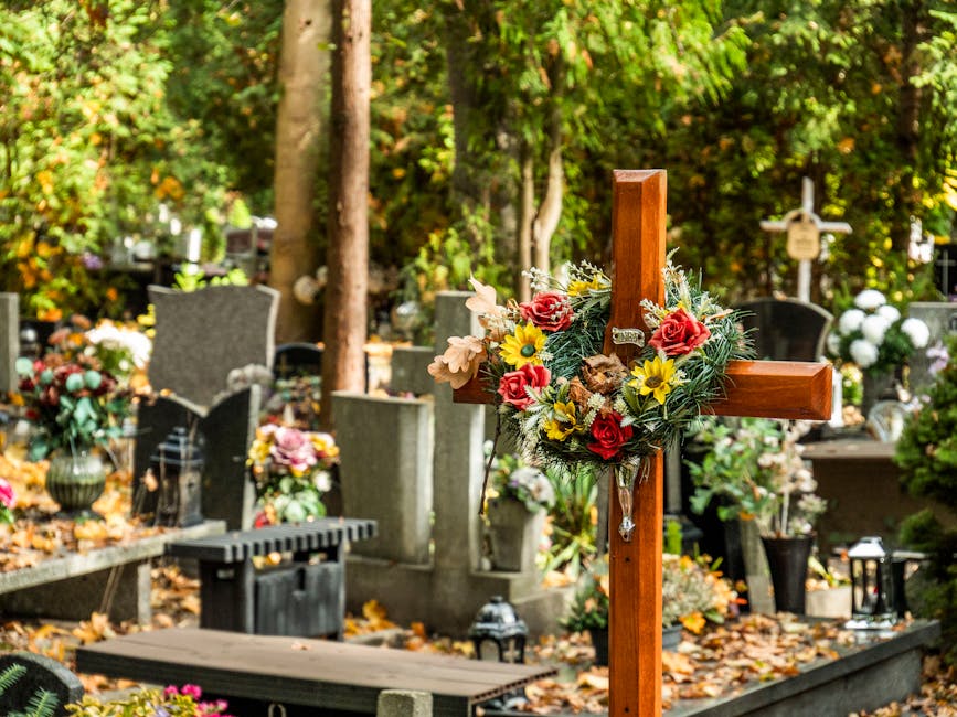 A wooden funeral cross adorned with a floral arrangement featuring pink roses, yellow sunflowers, and white lilies, placed among gravestones at Danson Park cemetery. The flowers appear fresh with deta