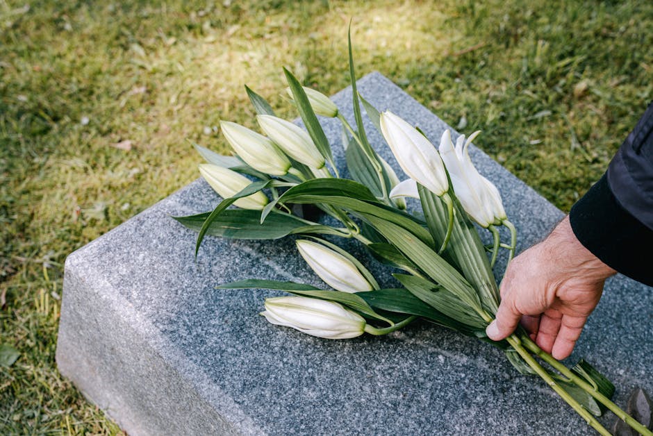 A person’s hand is gently placing a bouquet of white lilies with long, green leaves onto a grey stone memorial. The lilies are fresh, with closed buds and open blooms, wrapped in a simple green stem
